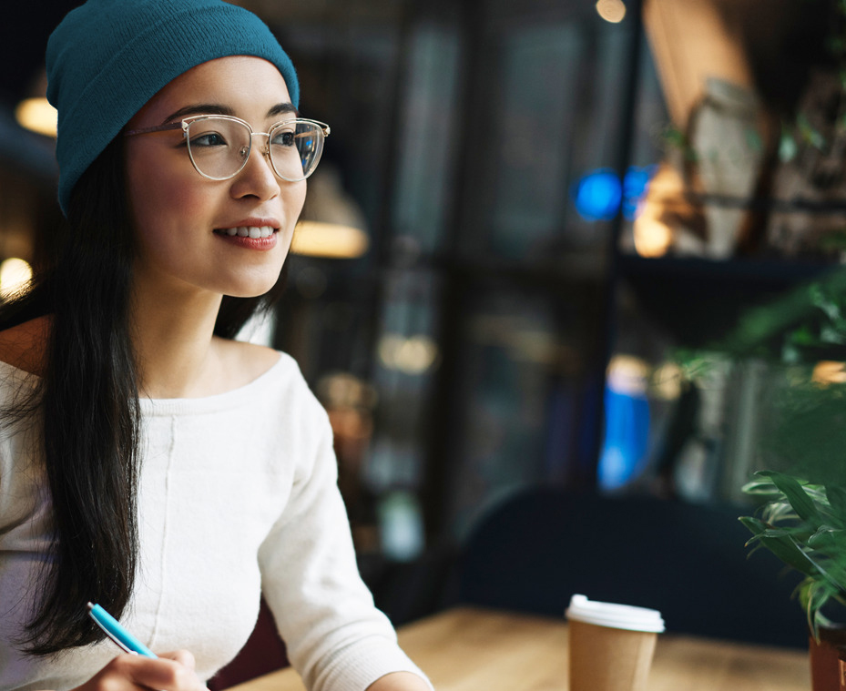 Woman in beanie and glasses in a coffee shop smiling while writing