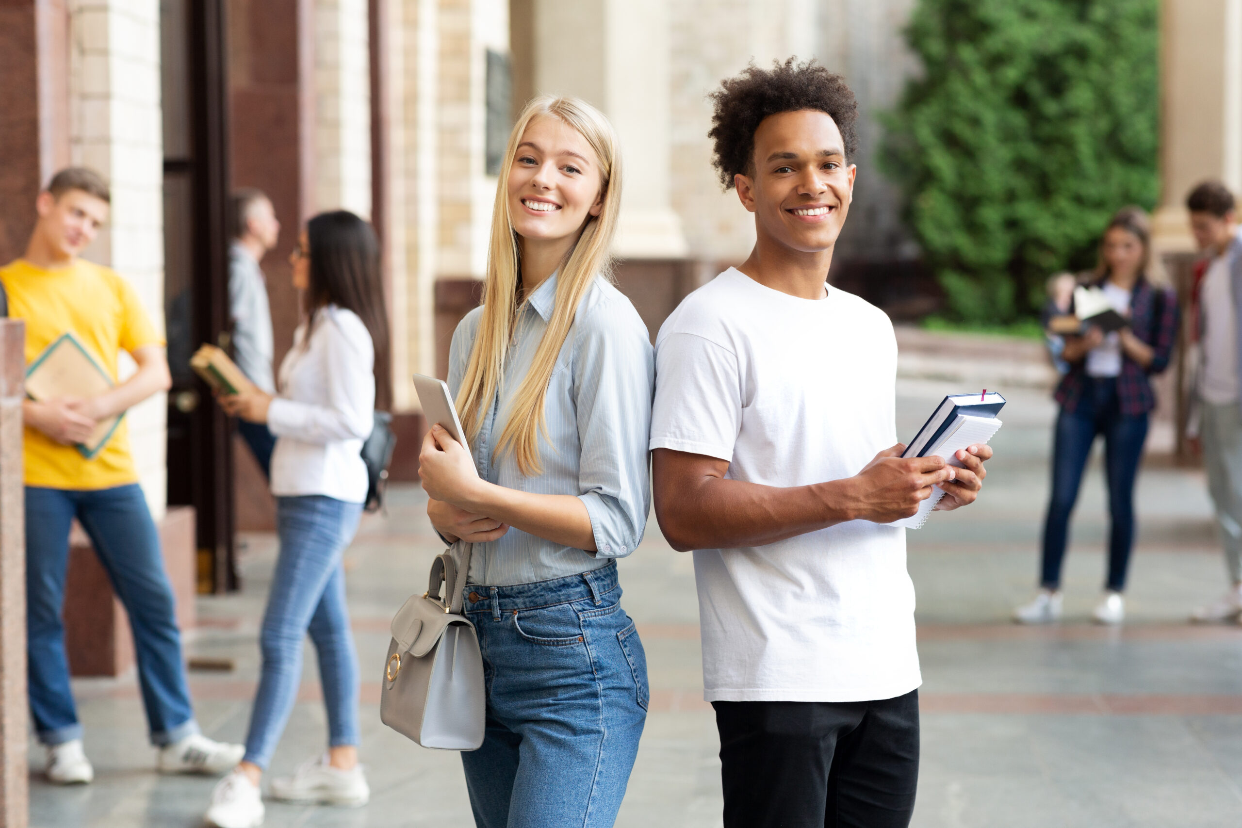 two students standing besides each other holding books in hand and similing